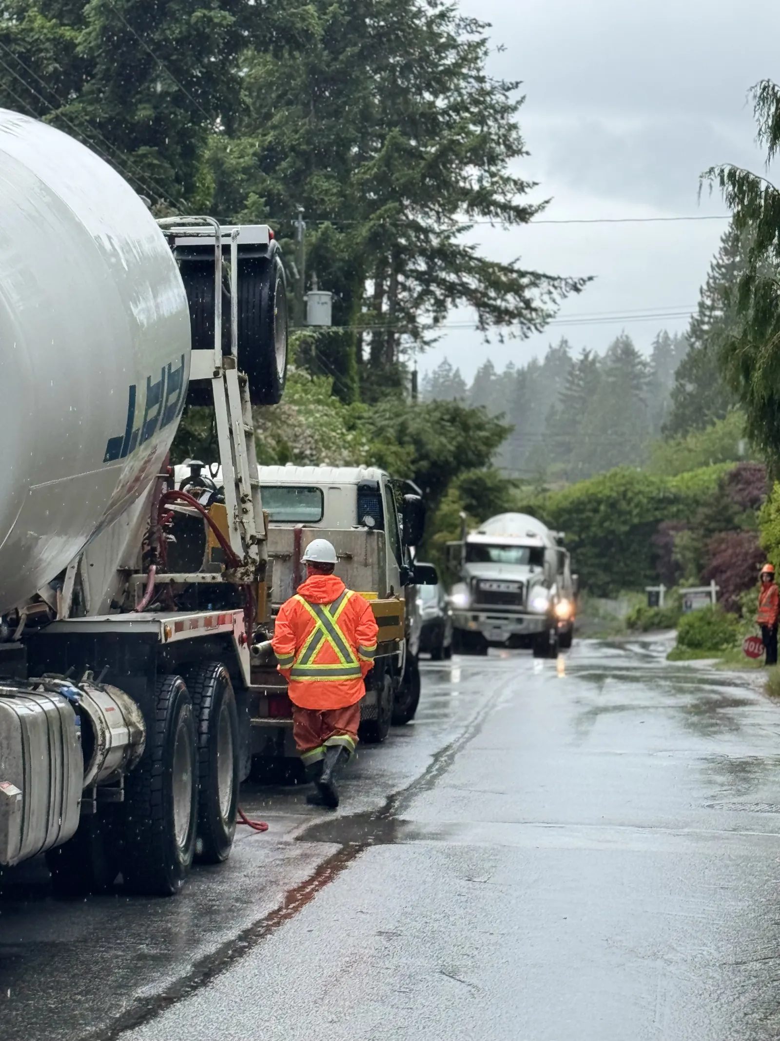 Concrete mixer trucks lined up on a wet, tree-lined North Shore street during a rainy pour, with a flagger in orange rain gear directing traffic