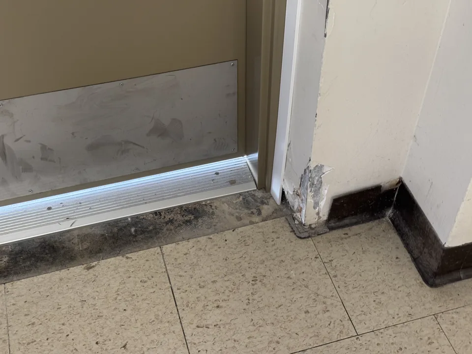 Water-damaged baseboard and flooring in a strata corridor, the interior consequence of an envelope or membrane left uninspected