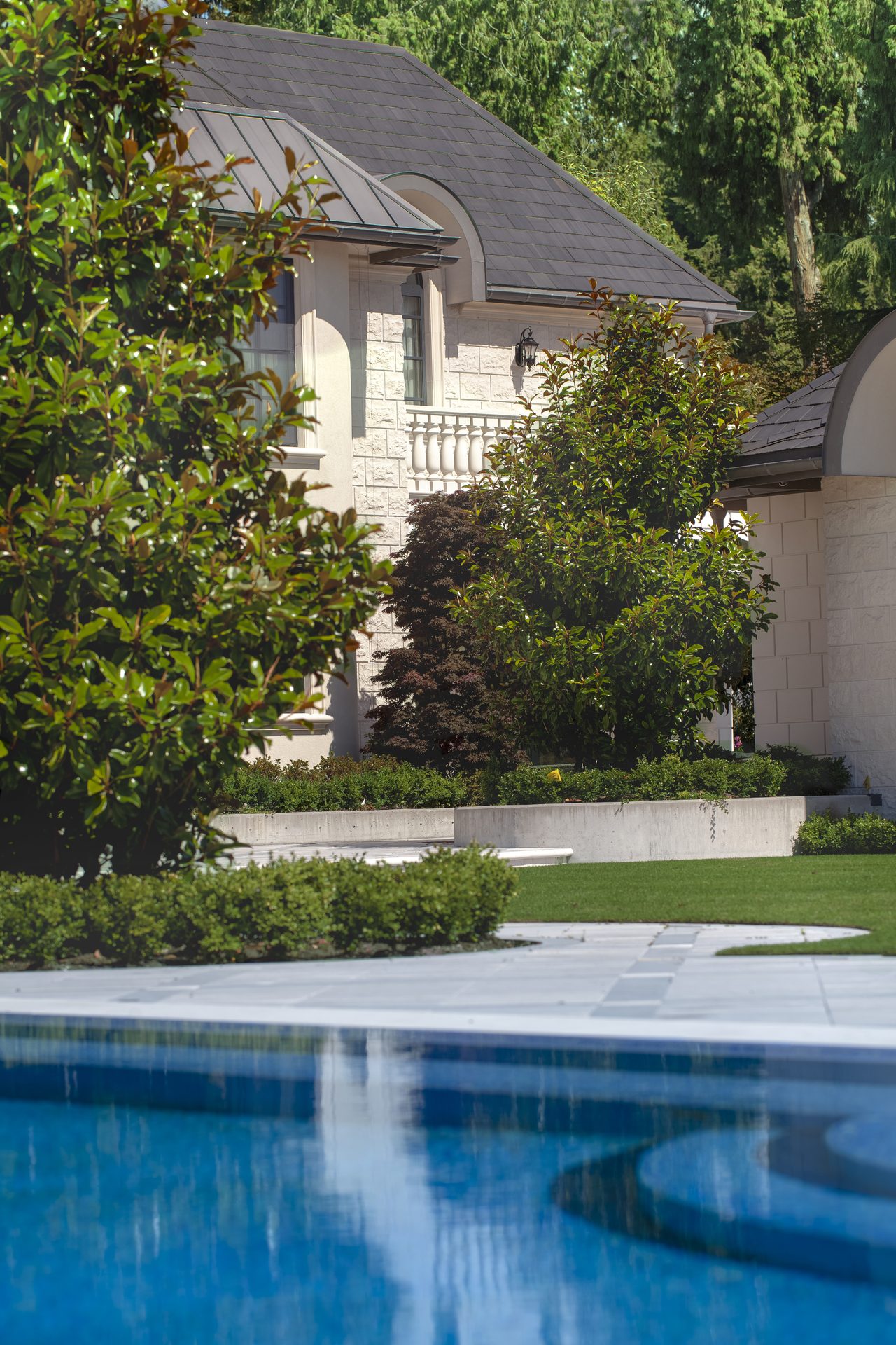 Ambleside Palace pool edge view toward the stone pavilion wing and mature landscaping in West Vancouver