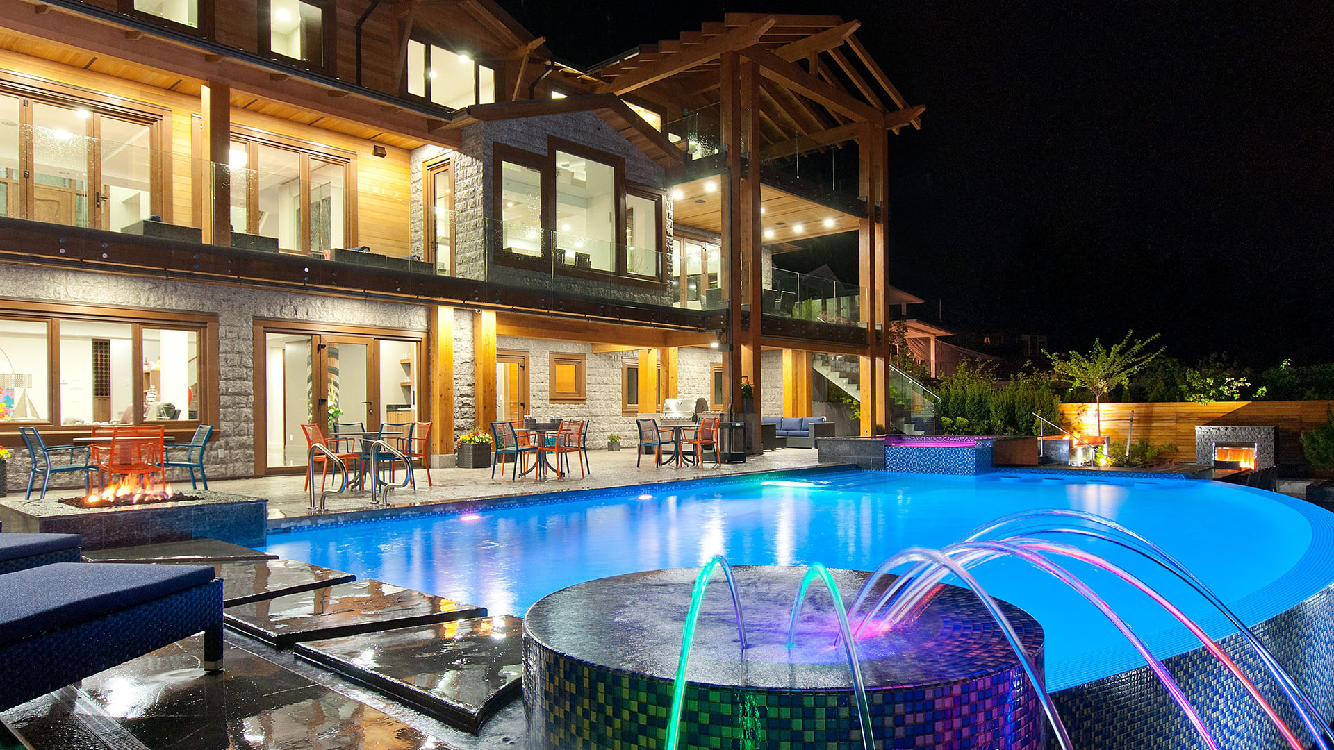 Night view of Millstream British Properties luxury pool terrace with illuminated waterfall jets fire features timber pergola and granite facade West Vancouver