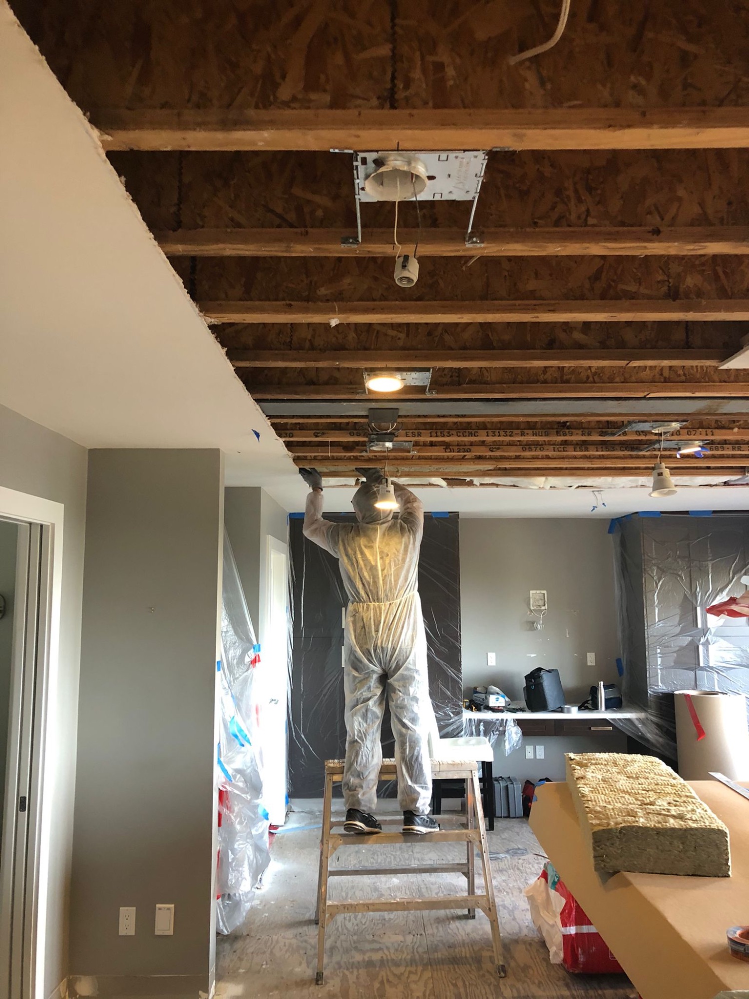 Worker in protective coveralls installing ceiling drywall and light fixtures at Newdale Court with exposed joists and mineral wool insulation visible