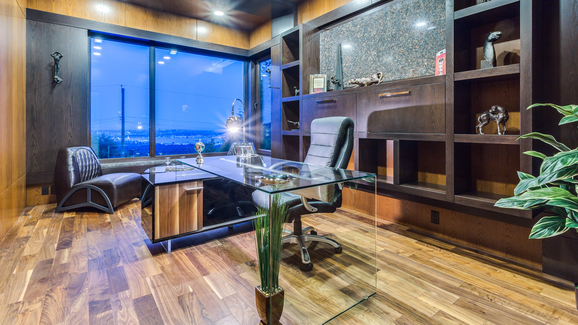 Walnut-panelled home office with built-in shelving glass desk and city views at twilight West Coast Contemporary British Properties West Vancouver